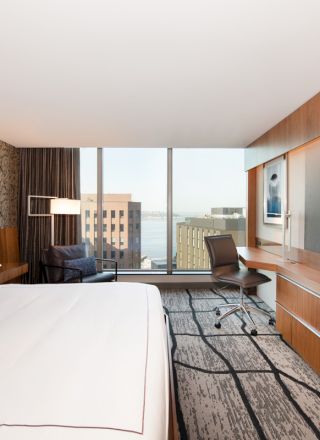A modern hotel room with a large bed, city view, desk, and flat-screen TV, featuring a contemporary design and patterned carpet.