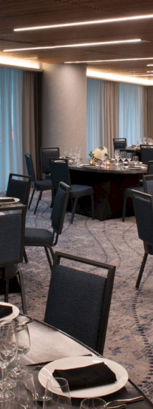 An elegant banquet room with round tables set for dining, featuring black chairs, dim lighting, and sheer blue curtains.