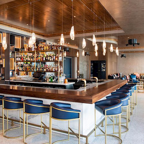 This image shows a modern bar with stylish seating, pendant lighting, and a well-stocked shelf of bottles in a spacious interior.
