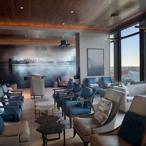 A modern lounge with plush seating, large windows offering a scenic view of water and a Ferris wheel, and wall art depicting a skyline.