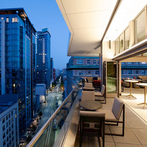 A modern urban balcony at dusk with sleek furniture overlooks a cityscape of tall glass buildings and streets below.