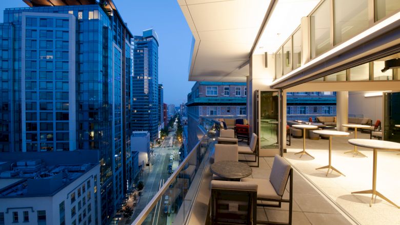 A modern cityscape at dusk with high-rise buildings, featuring a stylish balcony with seating and tables overlooking the street scene.
