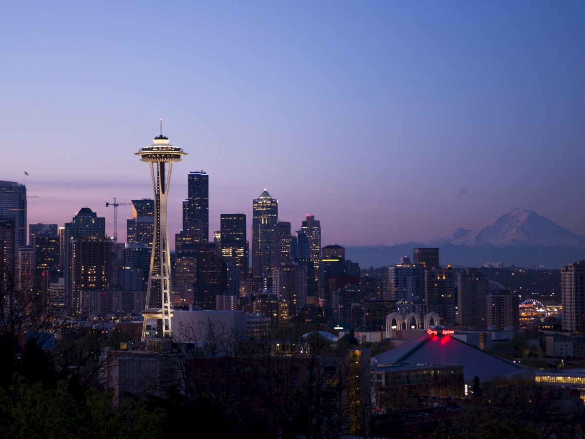 Seattle skyline at dusk with the Space Needle prominent, Mount Rainier in the distance, and city lights starting to illuminate.