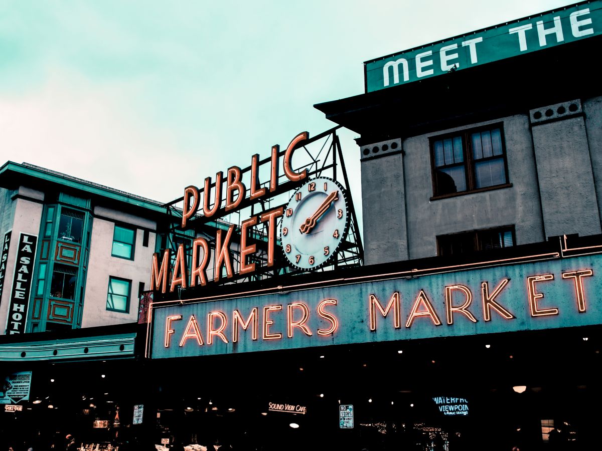 The image shows a public market sign reading "Farmers Market" with a neon design, attached to a building with an overcast sky.