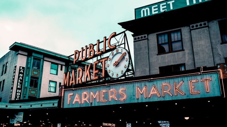 The image shows a public market sign reading "Farmers Market" with a neon design, attached to a building with an overcast sky.