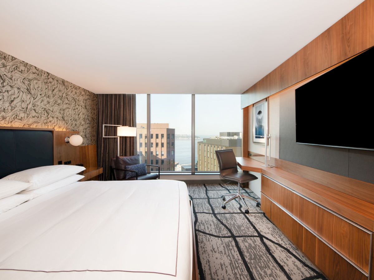 A modern hotel room with a large bed, wall-mounted TV, desk, and window view of buildings and water.