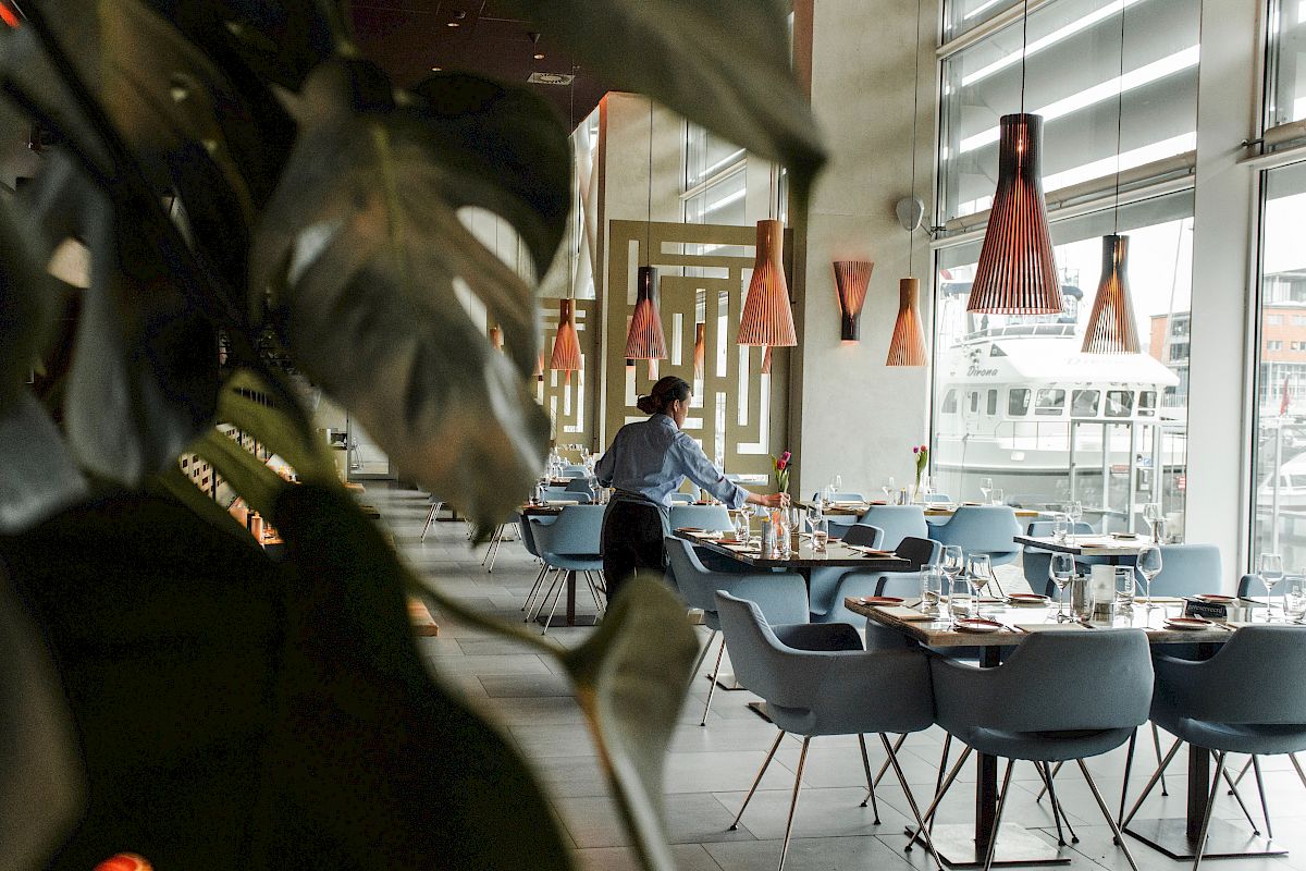 A person is setting tables in a modern restaurant with stylish lighting and large windows, surrounded by plants.