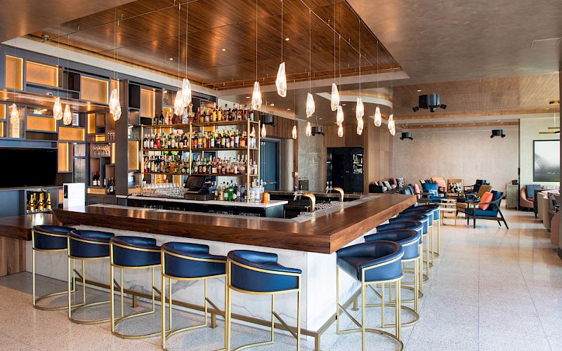 The image shows a modern bar with stylish lighting, a stocked back bar, blue cushioned stools, and a seating area in the background.