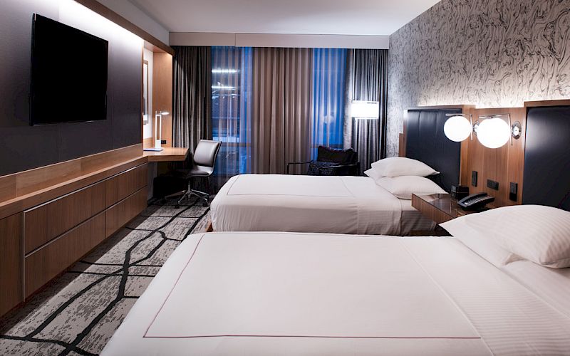 A modern hotel room features two beds, a TV on the wall, a desk with a chair, and stylish lighting and decor.