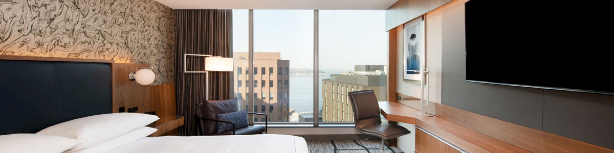 A modern hotel room with a large bed, TV, desk, chair, and window view of city buildings.